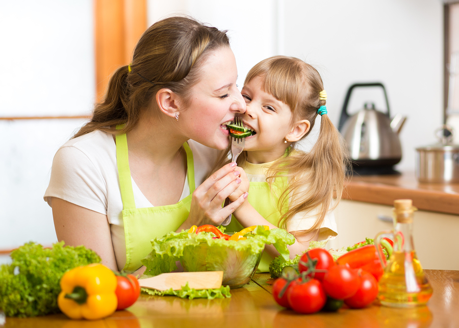 Mother Feeding Kid Vegetables In Kitchen